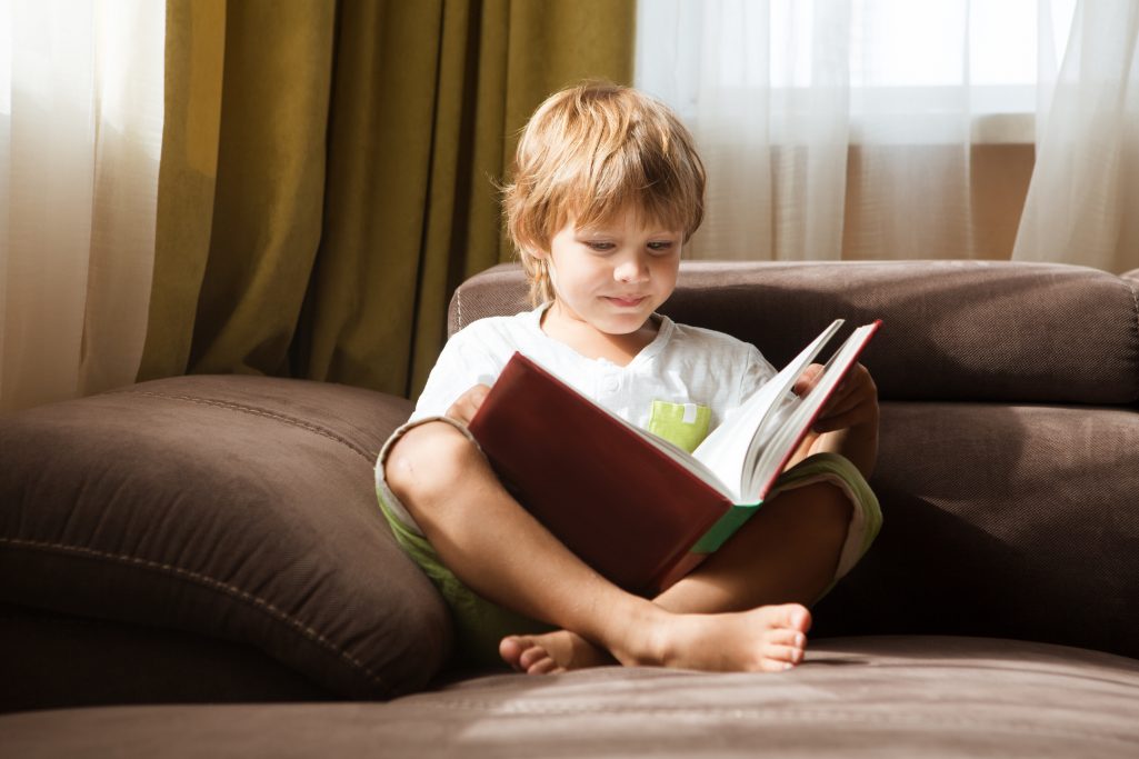 Cute little boy is reading book sitting on a sofa at home